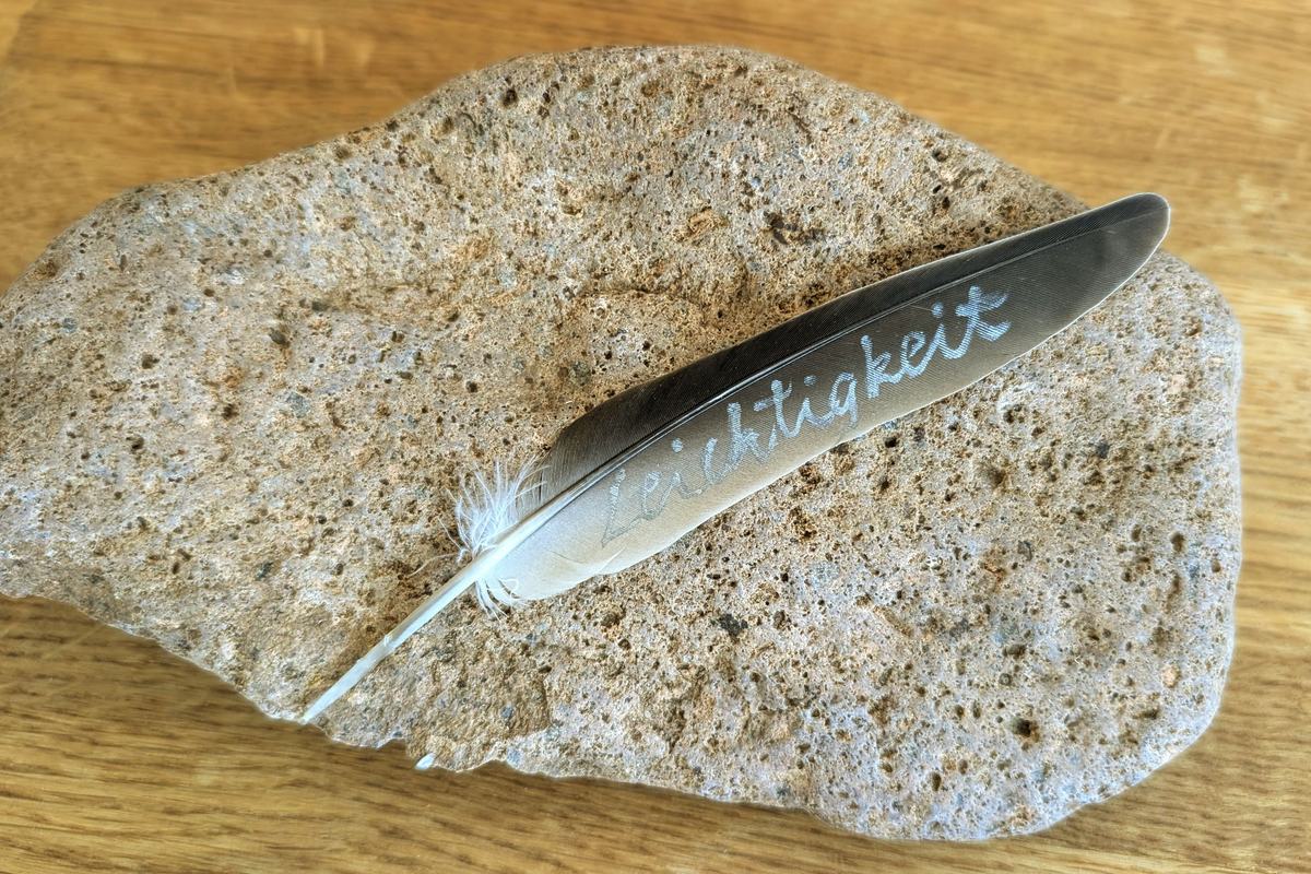 Eine helle Vogelfeder liegt auf einem schweren grauen Stein. Auf der Feder steht in Handschrift das Wort Leichtigkeit.