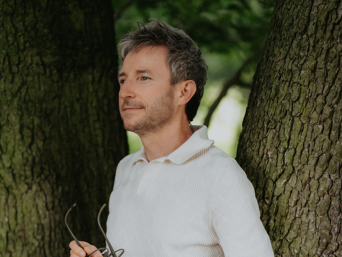Tobias Weise lehnt an einem alten Baumstamm und blickt ruhig zur Seite. In der Hand hält er seine Brille, hinter ihm grünes Sommerlicht.